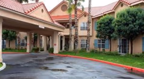 Exterior view of a senior living facility with a covered entrance, beige stucco walls, red tile roof, palm trees, and green landscaping along the driveway.