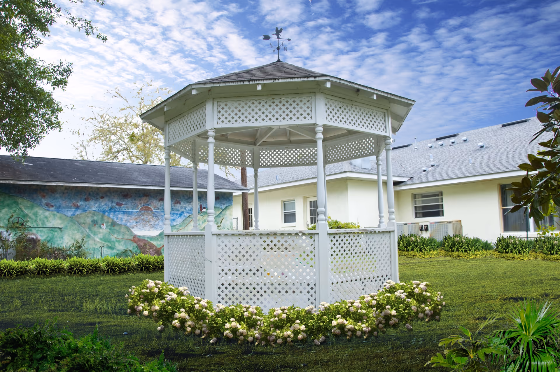 A white wooden gazebo with lattice panels stands in a grassy garden area surrounded by flowering plants. Behind the gazebo, there is a building with light-colored walls and a mural painted on one side. The sky is partly cloudy with blue patches visible.