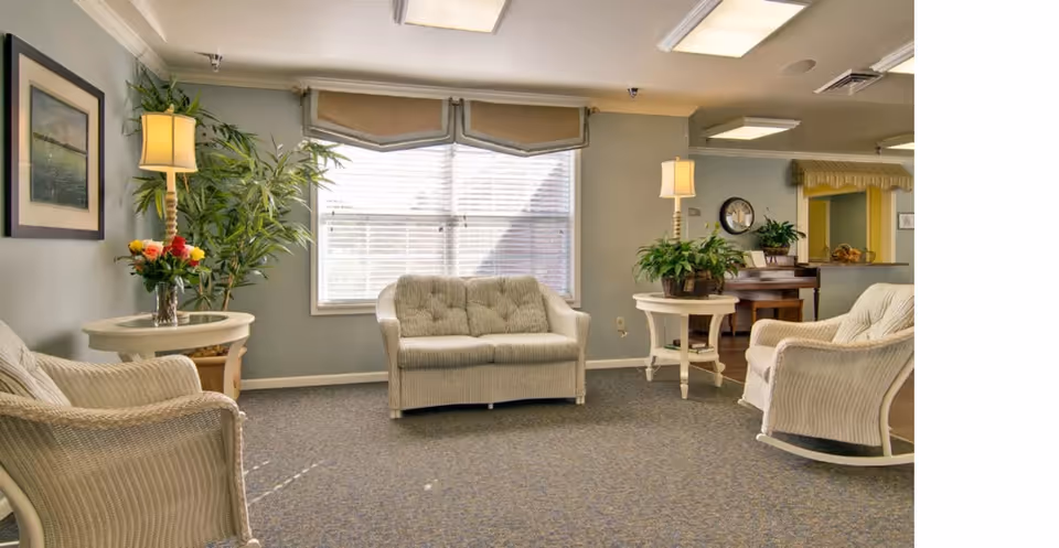 A cozy living room area with beige wicker furniture including a loveseat and two armchairs. There are two round side tables with lamps and plants on them. A large window with blinds and a valance lets in natural light. The walls are painted light green, and there is a framed picture on the left wall. In the background, a piano and a clock are visible.