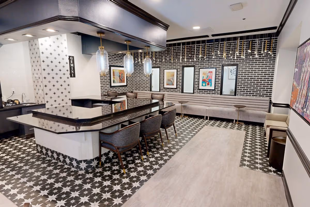 Modern common area with a black and white star-patterned tile floor, a large black countertop island with three chairs, pendant lights hanging above, a long gray cushioned bench along a black brick wall with framed artwork and mirrors, and additional seating including a beige chair and small tables.