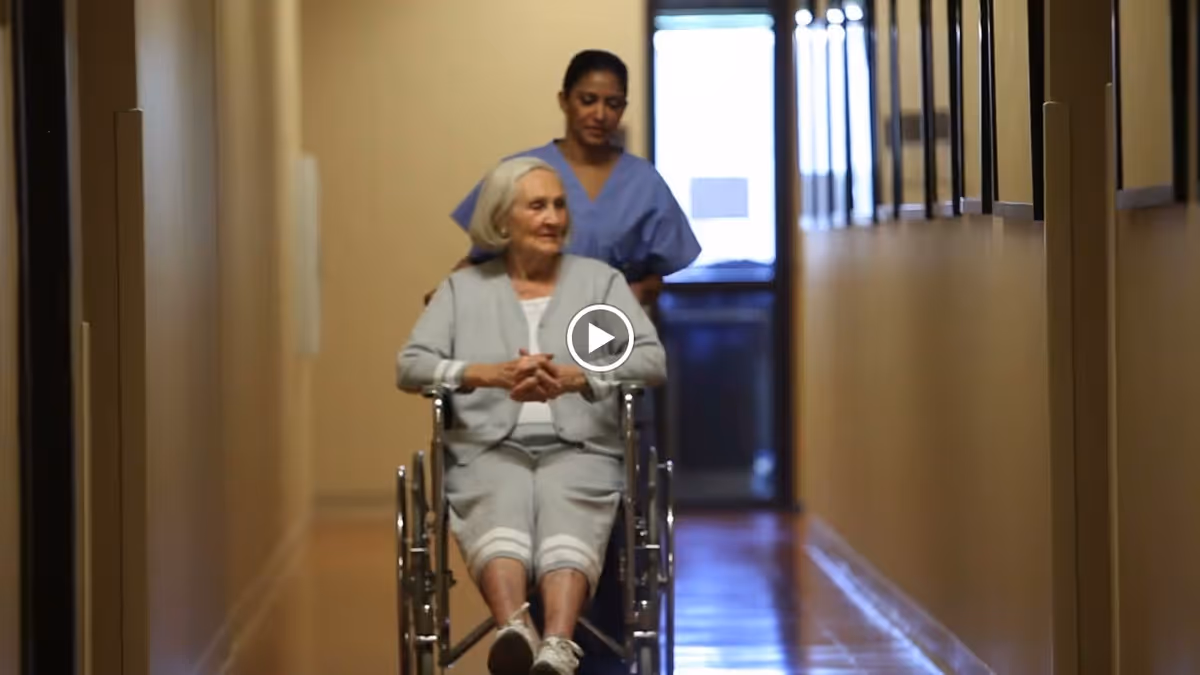 An elderly woman in a wheelchair being pushed down a hallway by a caregiver wearing blue scrubs inside a senior living facility.