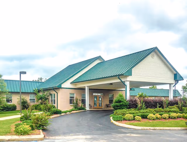 Front view of a single-story brick building with green metal roofs, a covered porte-cochere, driveway, and landscaped shrubs.