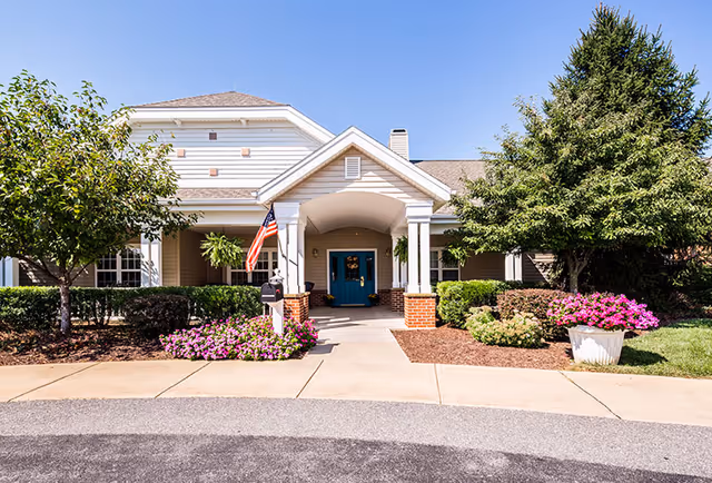 Front exterior view of Somerford House And Place Hagerstown showing a single-story building with a covered entrance, white columns, a blue door, an American flag, and landscaped bushes and flowers under a clear blue sky.