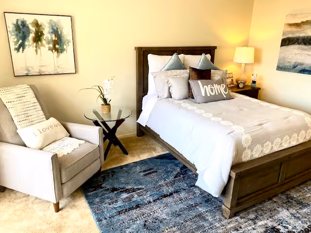 Bright furnished bedroom with a wooden bed and decorative pillows (one reading 'home'), an upholstered armchair with a throw, glass side table, lamp, and patterned rug.
