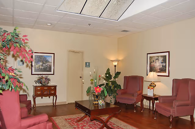 A cozy sitting area with four red upholstered armchairs arranged around a dark wooden coffee table with a floral centerpiece. The room has beige walls with two framed paintings, a small wooden side table with a lamp, a floor lamp in the corner, and a door with a restroom sign. There are also some potted plants and a red patterned rug on the wooden floor.