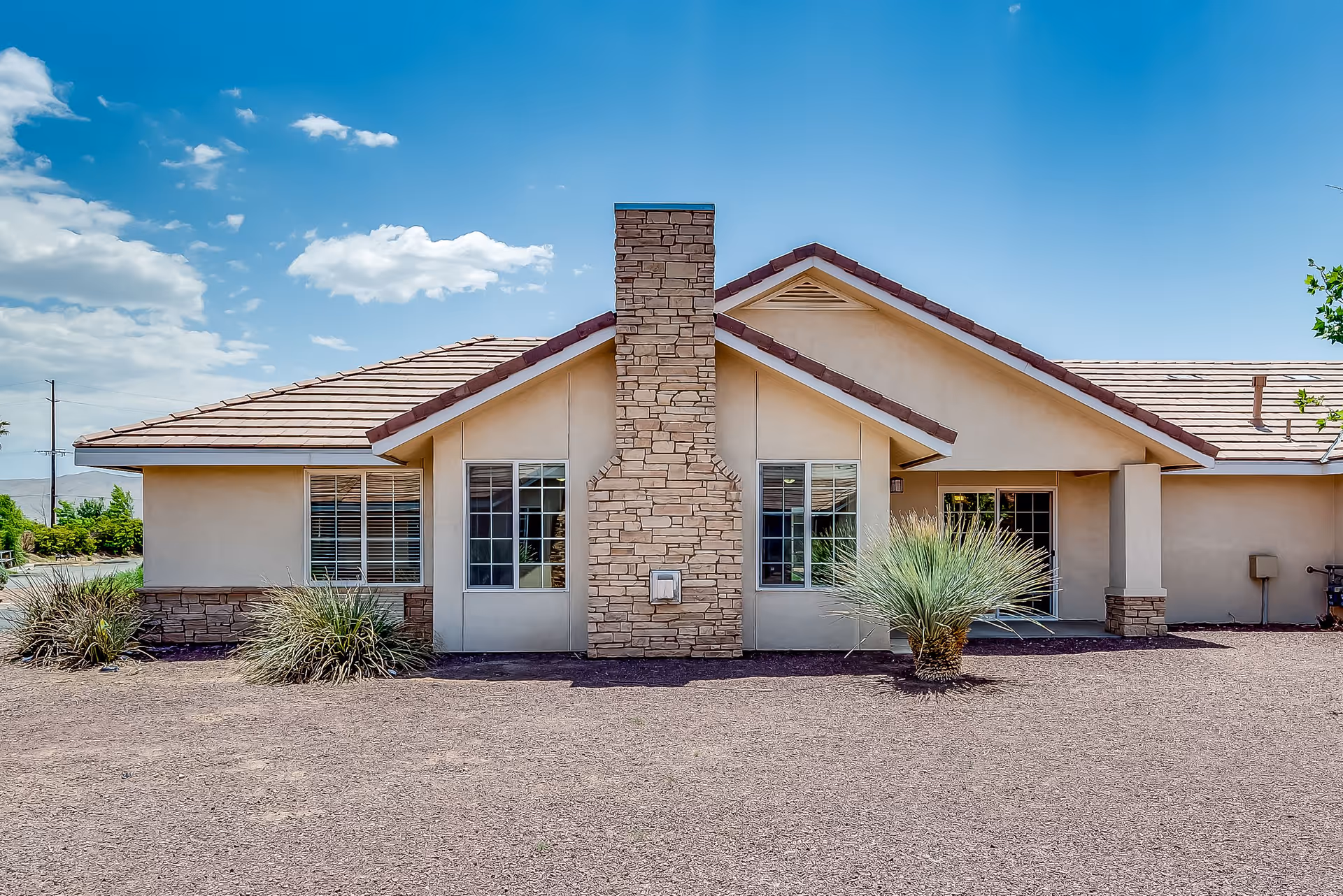 Exterior view of a single-story building with beige walls, a stone chimney in the center, and a tiled roof under a blue sky with some clouds. There are two windows on either side of the chimney and a door on the right side. Desert landscaping with small bushes and gravel surrounds the building.