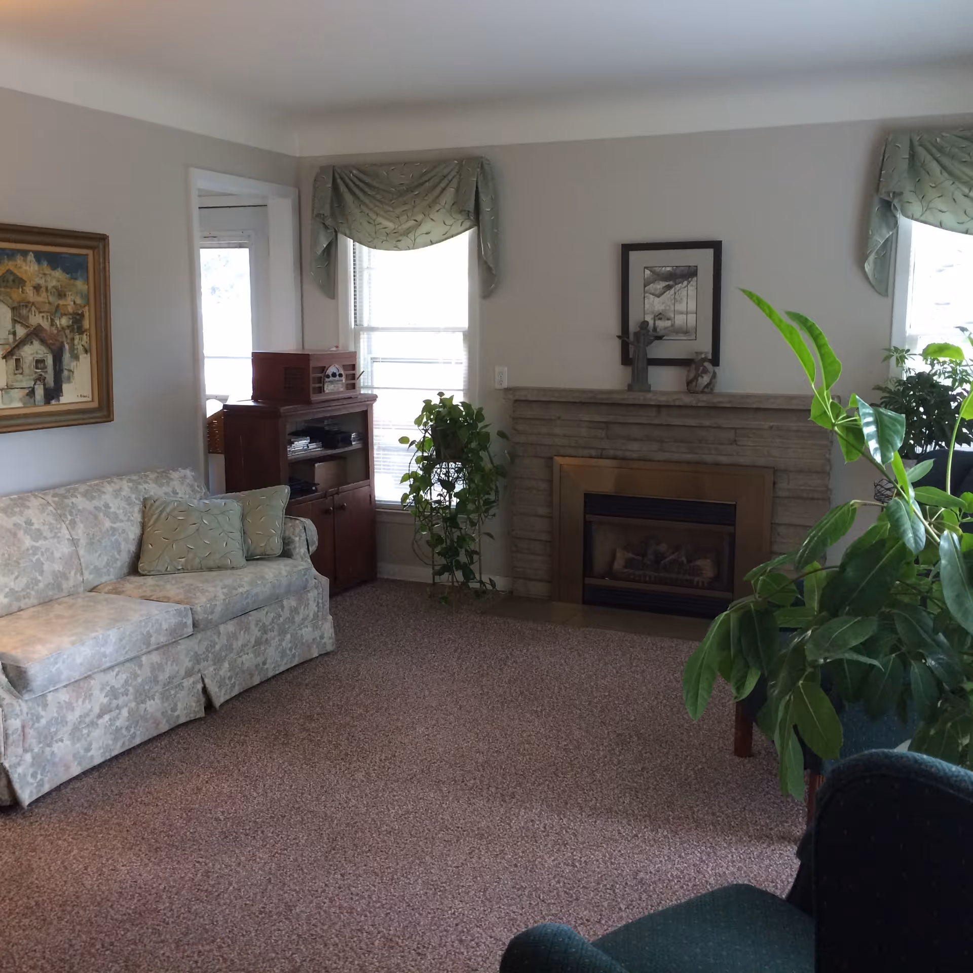 A cozy living room with a floral-patterned sofa with two matching pillows, a stone fireplace with a framed picture and decorative items on the mantel, two windows with green valances, a wooden cabinet with a vintage radio on top, and several green potted plants.