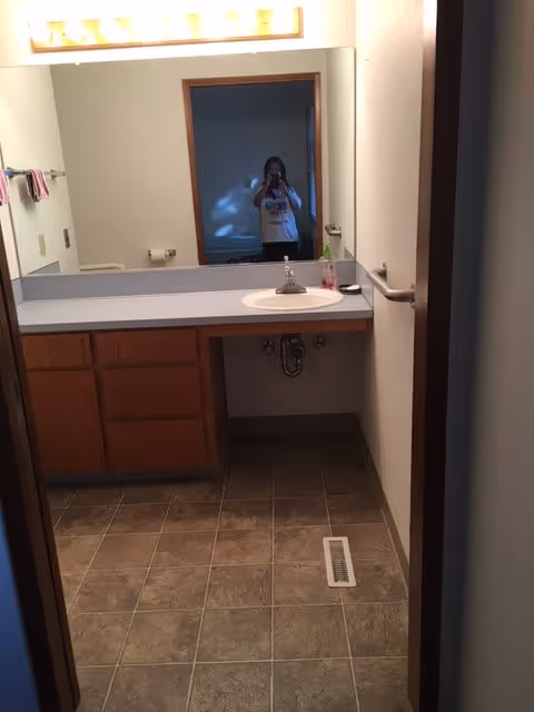 A bathroom with a large mirror above a countertop with a sink. There are wooden cabinets below the countertop. A person is visible in the mirror taking the photo. The floor is tiled, and there is a vent on the floor. A towel rack with towels is mounted on the left wall.