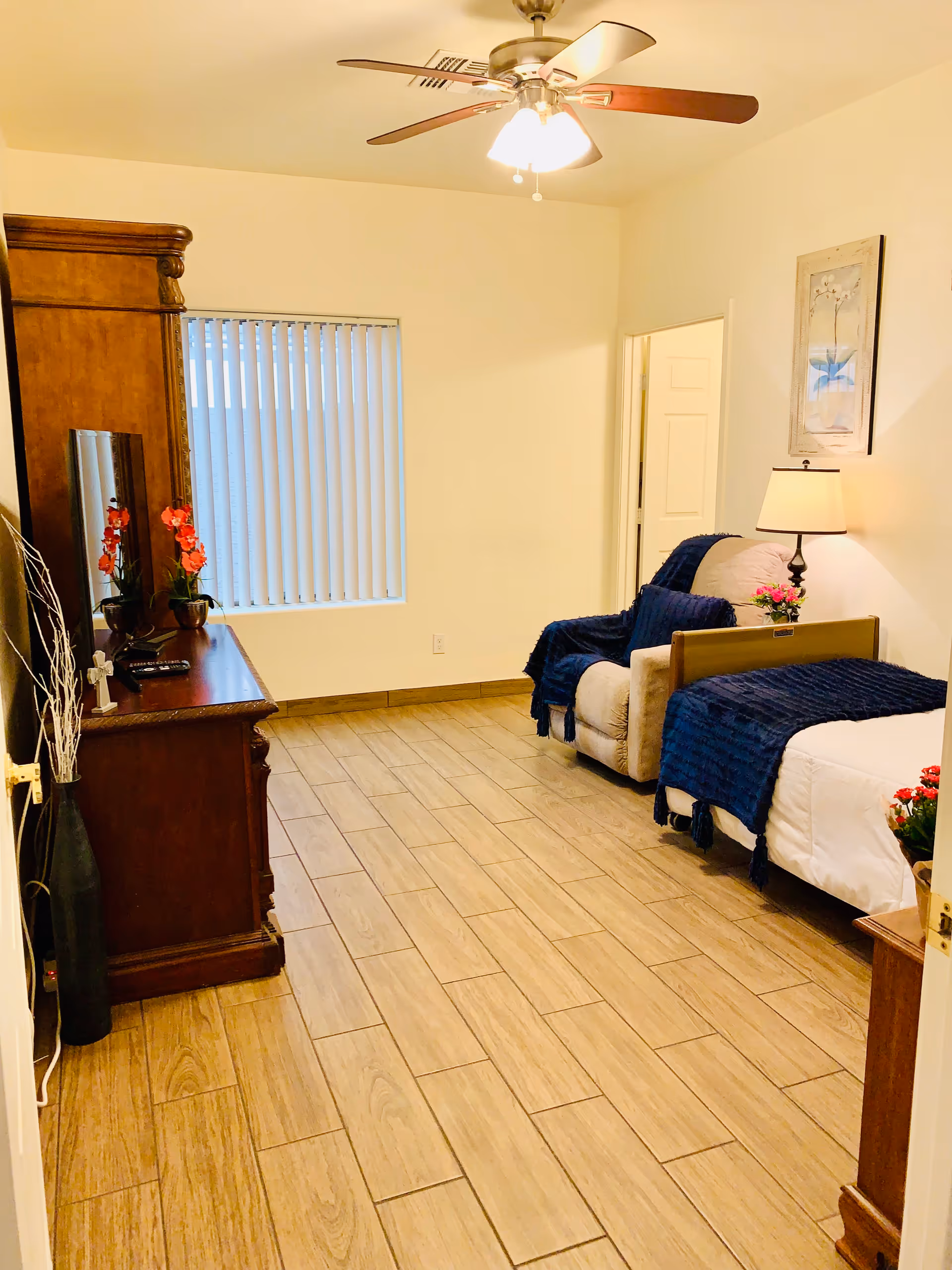 Well-lit bedroom with a single bed, upholstered recliner, wooden dresser, ceiling fan, and tiled floor.