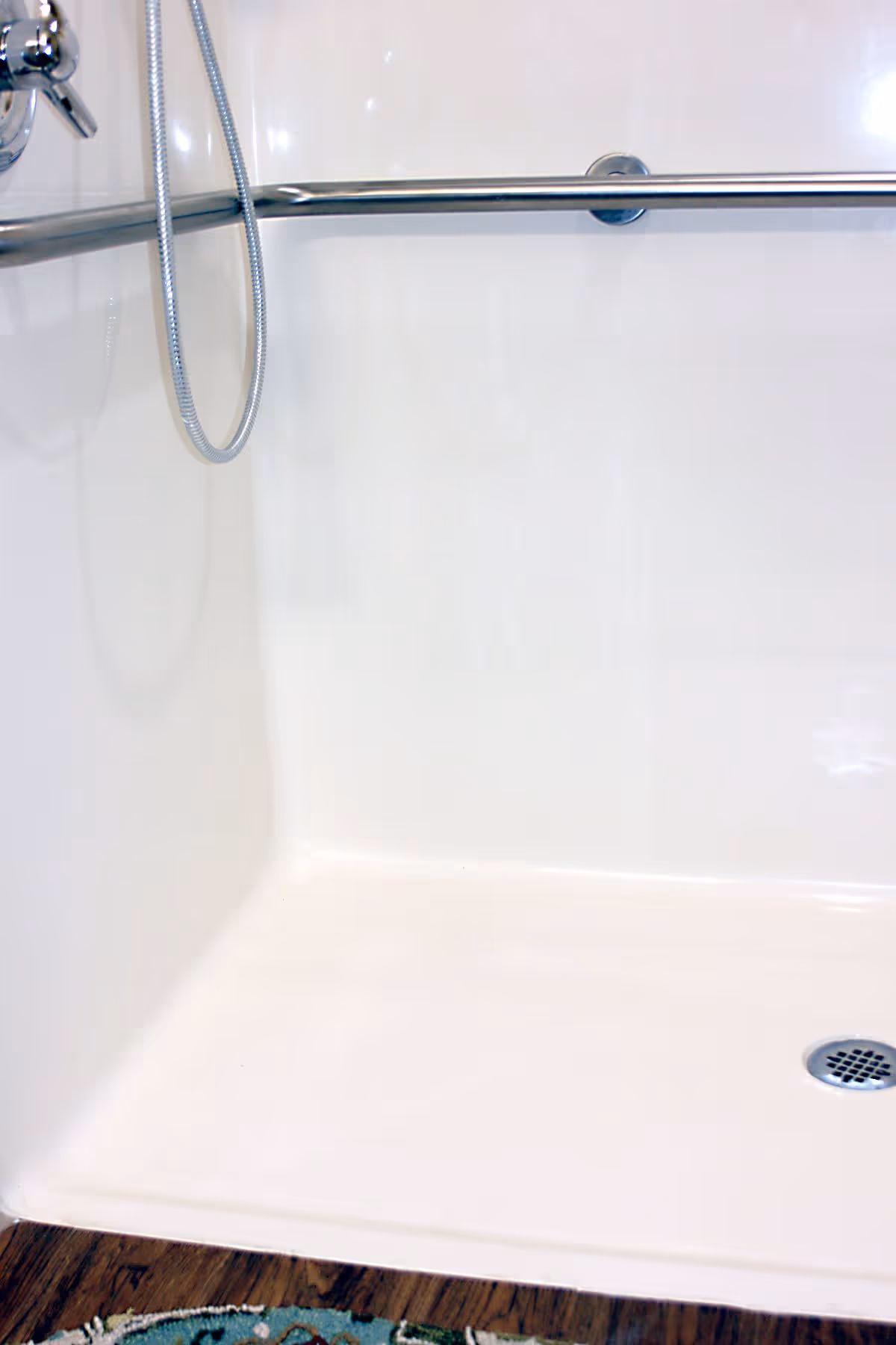 Close-up view of a white shower area with a silver grab bar mounted on the wall, a handheld showerhead with a flexible hose, and a drain on the shower floor. The floor outside the shower is wooden with a small patterned rug partially visible.