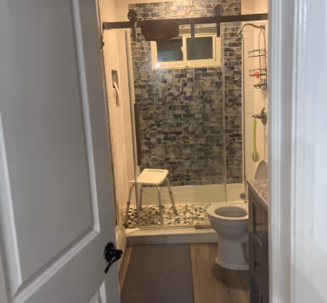 View through an open door into a bathroom with a toilet and vanity on the right and a walk-in shower with blue mosaic tile, pebble floor, and a shower stool.