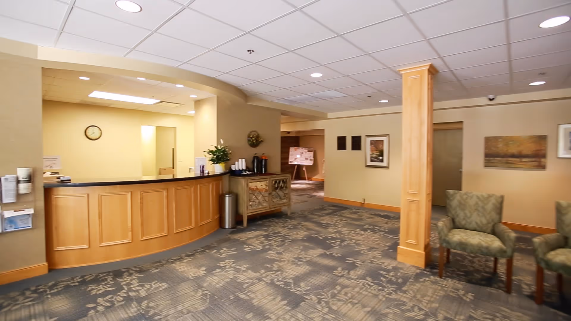 Reception area of a senior living community with a wooden front desk, a small table with coffee supplies, patterned carpet, beige walls, two upholstered chairs, and framed artwork on the walls.