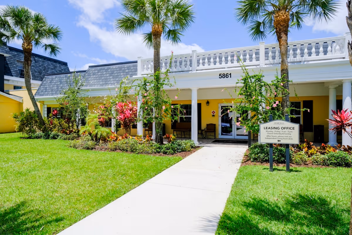 Exterior view of Volante Senior Living of Delray Beach leasing office building with a white walkway leading to the entrance, surrounded by green grass, palm trees, and colorful plants under a blue sky with some clouds.