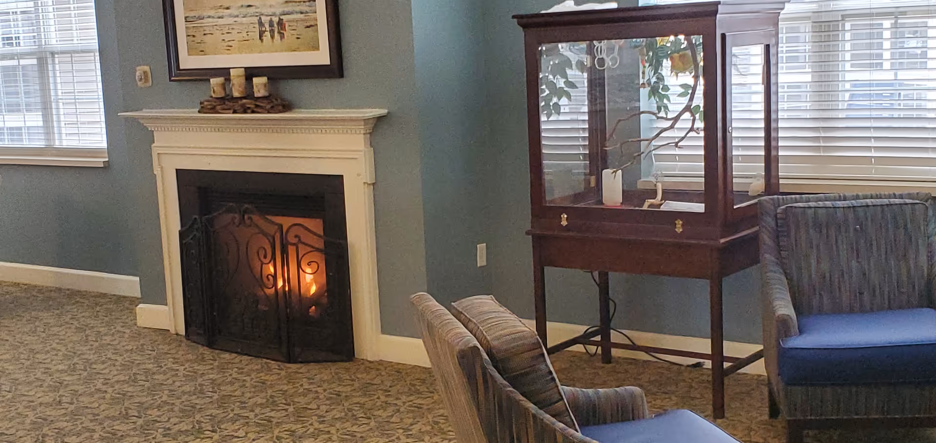 A cozy interior room with a lit fireplace featuring a decorative black metal screen. Above the fireplace is a framed painting and three candles on a wooden tray. To the right, there is a wooden display cabinet with glass panels and a plant inside. Two upholstered chairs with blue cushions are positioned near the cabinet. The room has blue walls, patterned carpet, and windows with white blinds.