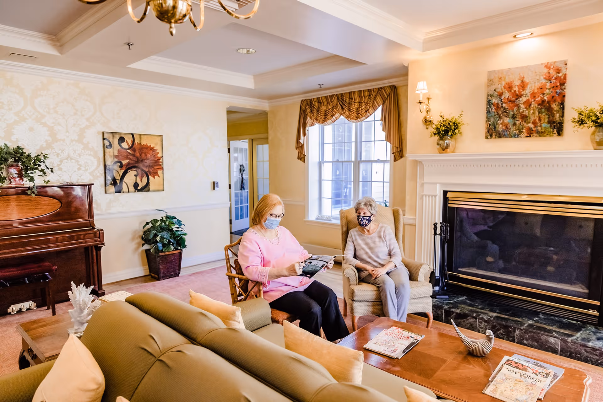 Two elderly women wearing face masks sitting in a cozy living room with a fireplace, a piano, and decorative plants. One woman is reading a magazine while the other is seated in an armchair. The room has warm lighting, patterned curtains, and artwork on the walls.