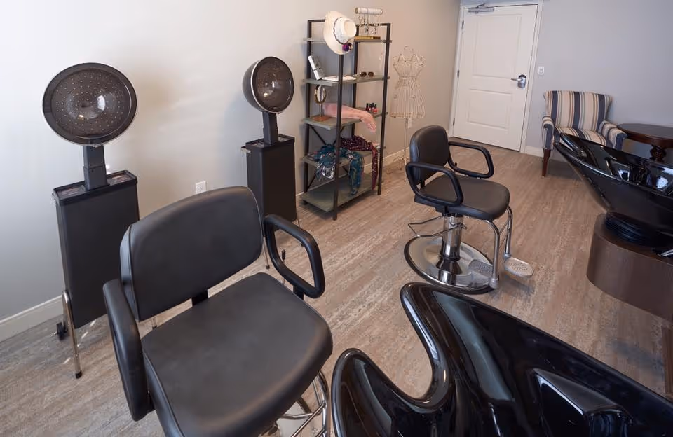 Small salon room with black styling chairs, hooded hair dryers, wash basins, and a shelving unit.
