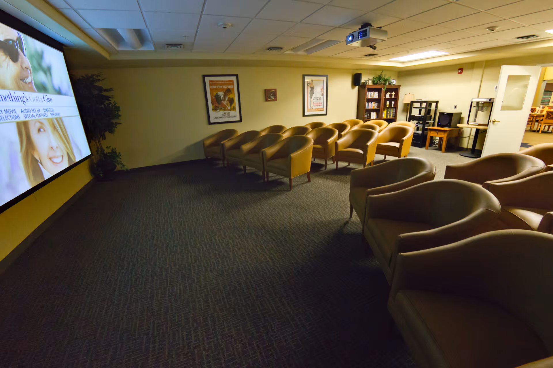 A small theater room with several rows of tan armchairs facing a large screen on the left wall. The room has a carpeted floor, framed posters on the back wall, a bookshelf with DVDs or books, and a projector mounted on the ceiling. There is a door on the right side leading to another room with tables and chairs.