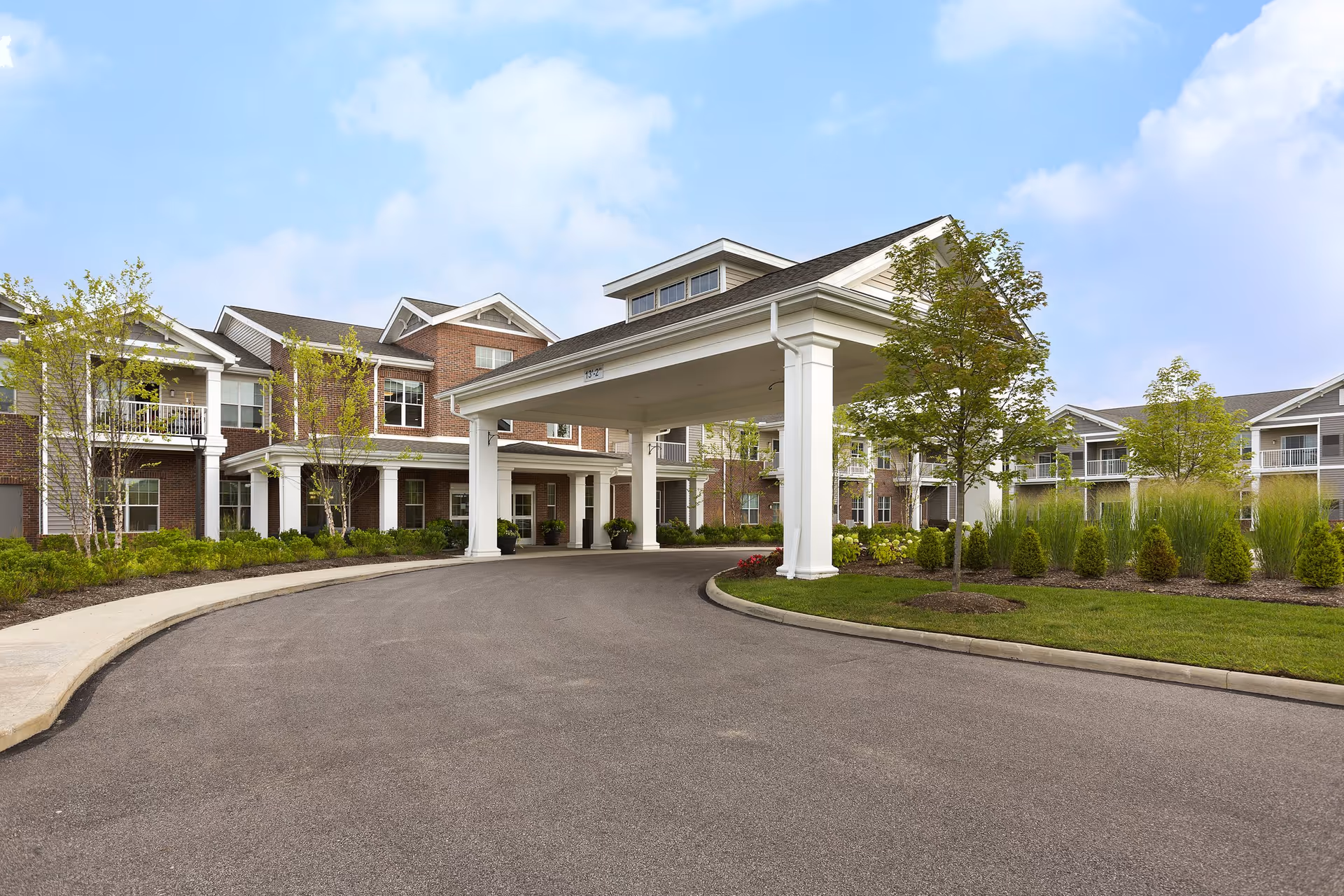 Exterior view of StoryPoint Medina senior living facility showing a large covered entrance with white pillars, a curved driveway, landscaped greenery, and a multi-story brick and siding building under a partly cloudy sky.