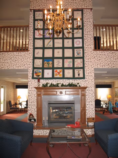 A cozy living room area with a wooden fireplace in the center, above which hangs a large quilt with various embroidered squares. A chandelier with lit candles hangs from the ceiling. Two blue upholstered chairs face a glass coffee table in front of the fireplace. The room has patterned wallpaper and a second-floor balcony railing visible above the fireplace. Lamps and additional seating are visible in the background.