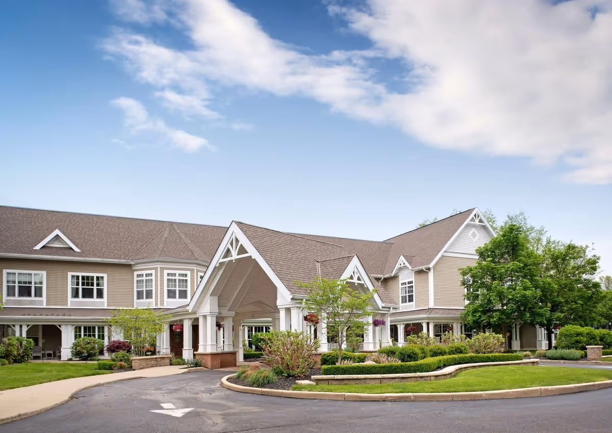 Exterior view of a senior living facility building with beige siding, white trim, and a covered entrance. The building is surrounded by well-maintained landscaping including bushes, trees, and hanging flower baskets. The sky is partly cloudy with blue patches.