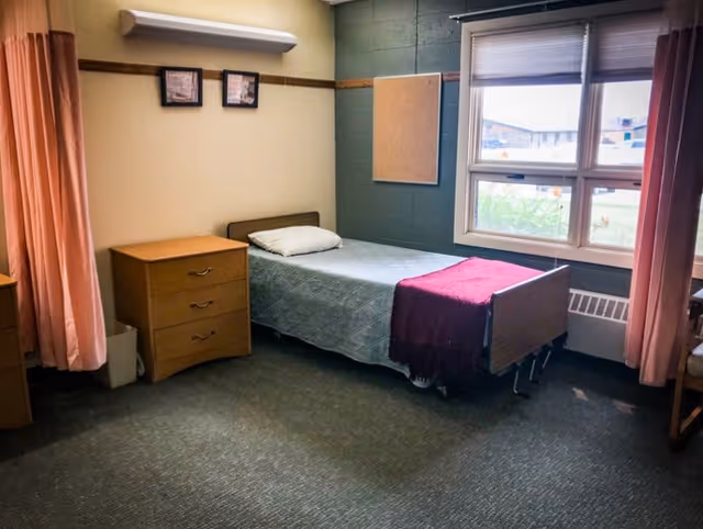 A simple bedroom in a senior living facility with a single bed covered with a light blue quilt and a dark red blanket at the foot. There is a wooden nightstand with three drawers next to the bed, a window with partially open blinds letting in natural light, and pink curtains on either side. The walls are painted beige and dark green, with a corkboard and two framed pictures hanging above the bed.