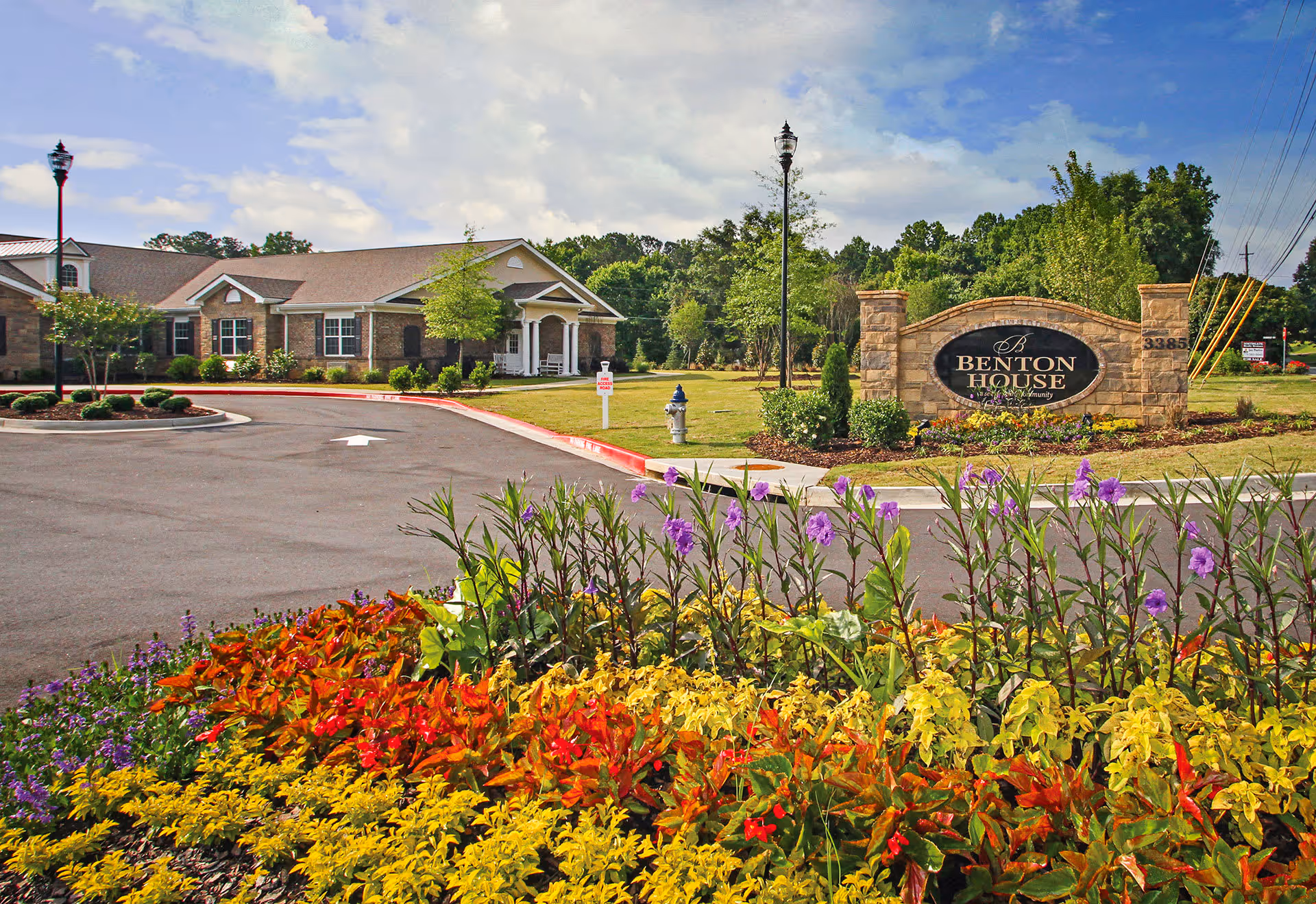 Entrance to Benton House of Woodstock senior living community with a stone sign, colorful flower bed in the foreground, a paved driveway, and a single-story brick building under a partly cloudy sky.