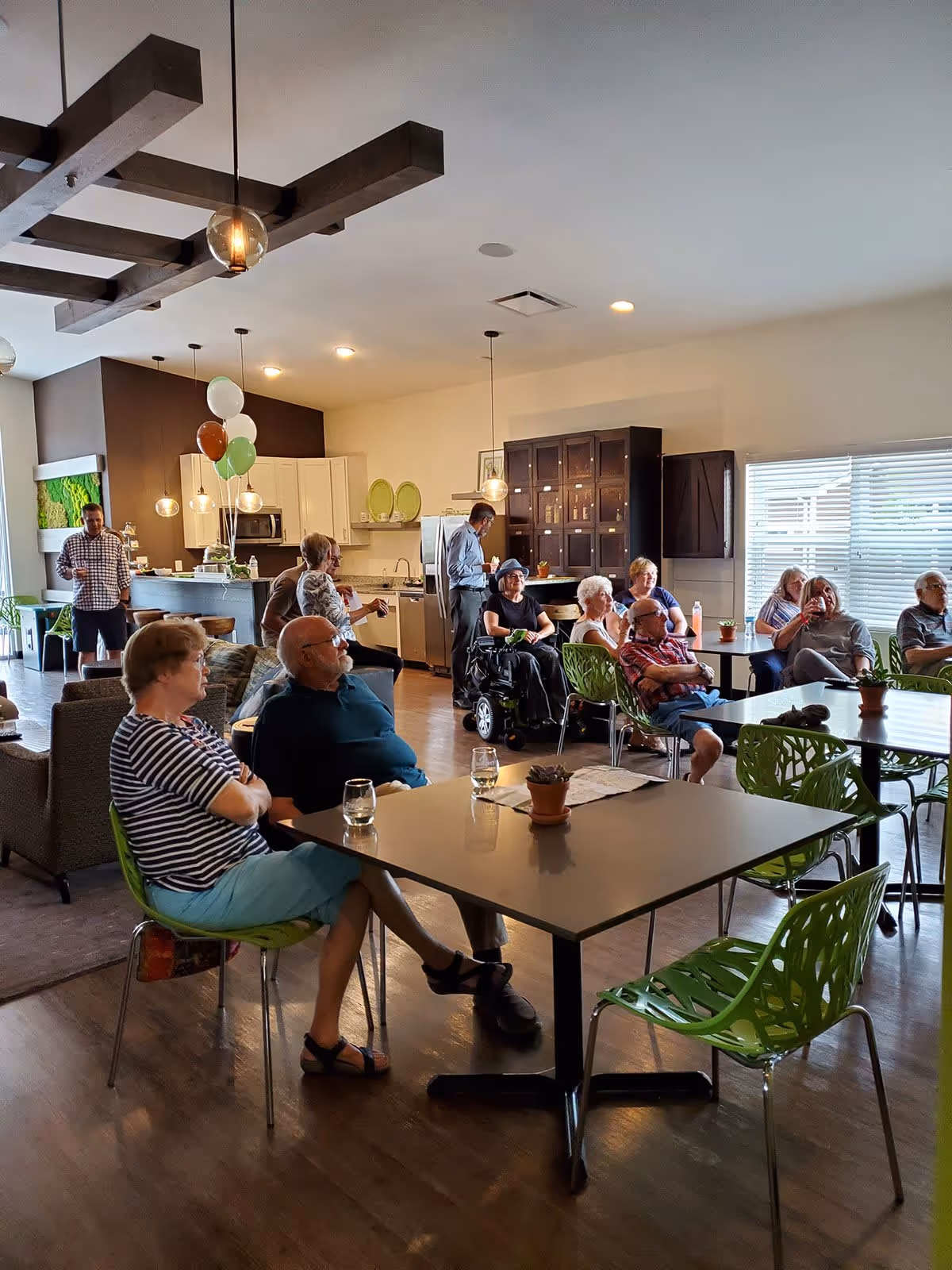 A group of elderly people sitting and socializing in a spacious, well-lit common area with modern furniture and a kitchen in the background. Some people are seated at tables with green chairs, while others are standing near the kitchen counter. The room has wooden floors, pendant lights, and large windows with blinds.