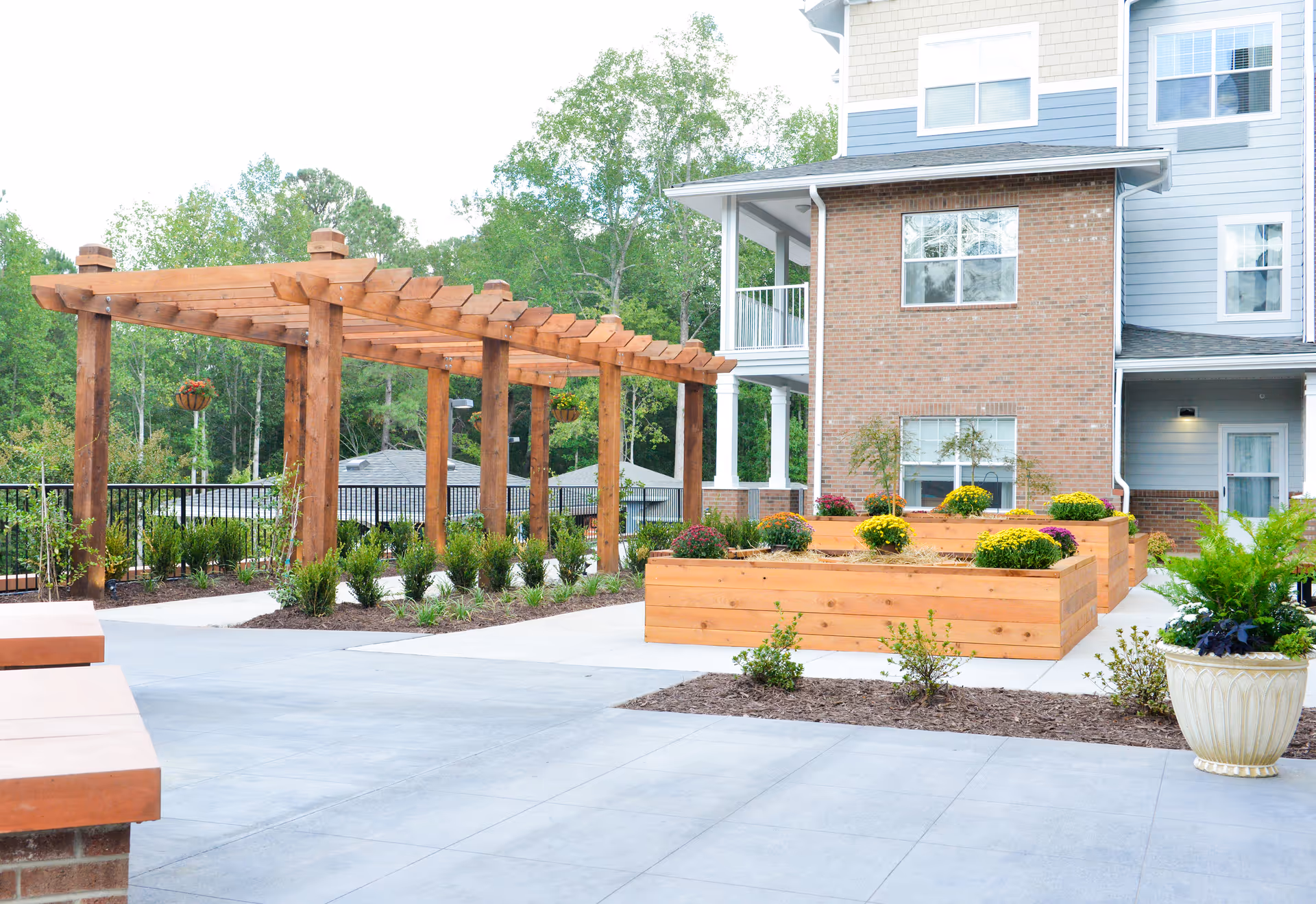 Outdoor courtyard with a wooden pergola, raised wooden planter boxes of flowers, and a multi-story brick-and-siding building in the background.