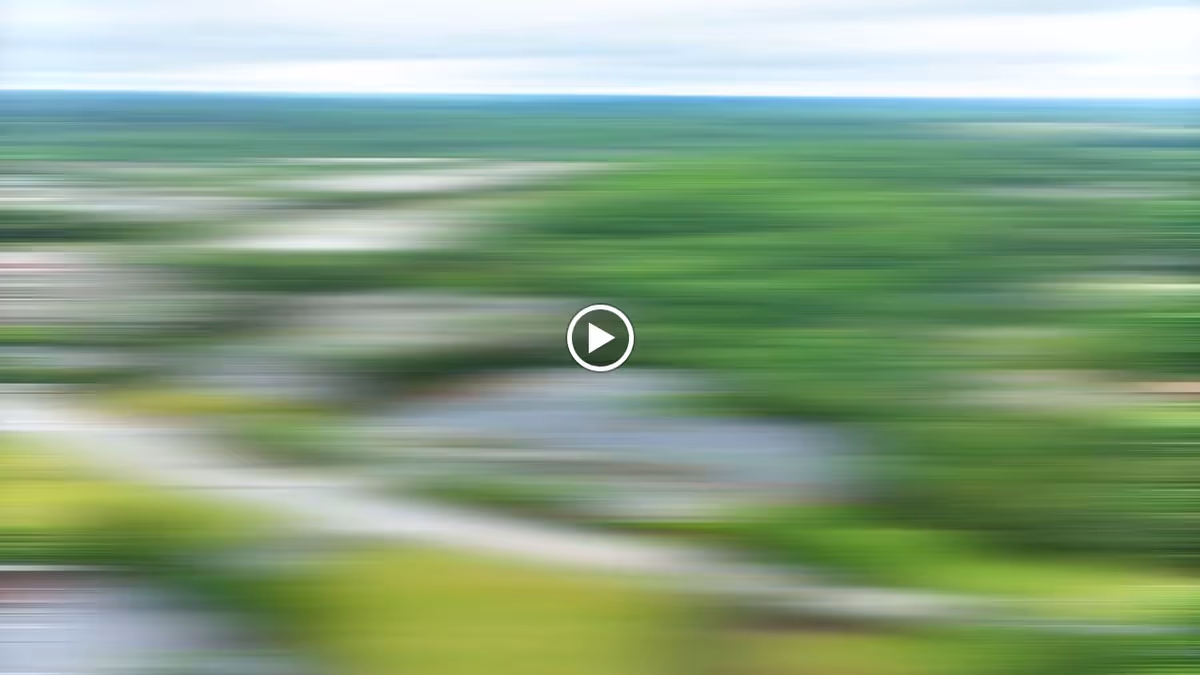 A blurred aerial view of a green landscape with trees, roads, and some buildings under a partly cloudy sky.