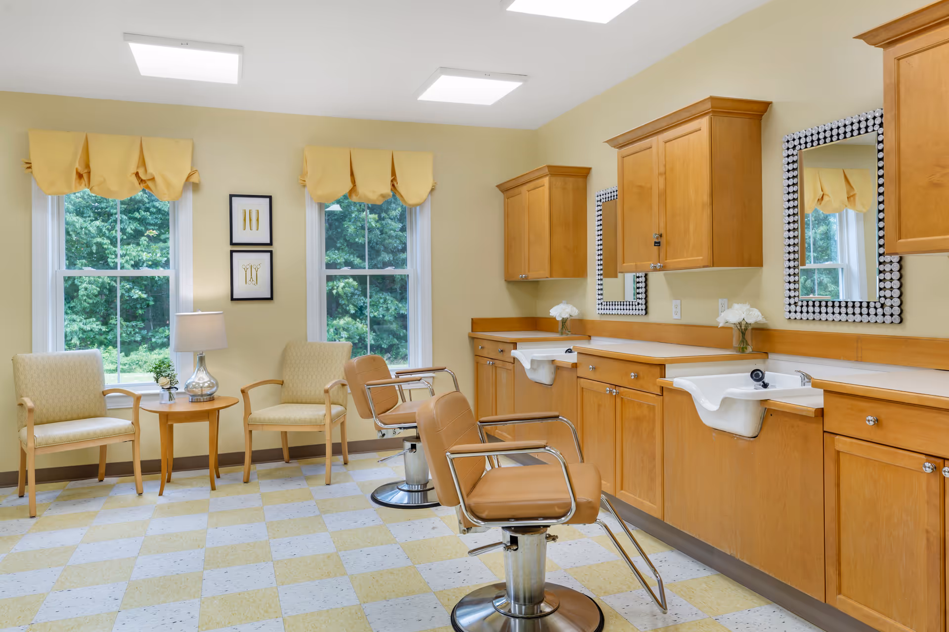A bright room with two large windows featuring yellow valances, two beige armchairs with a small round wooden table and lamp between them, and two tan salon chairs in front of a counter with sinks and mirrors framed with decorative black and white patterns. The room has light yellow walls and a checkered yellow and white floor.
