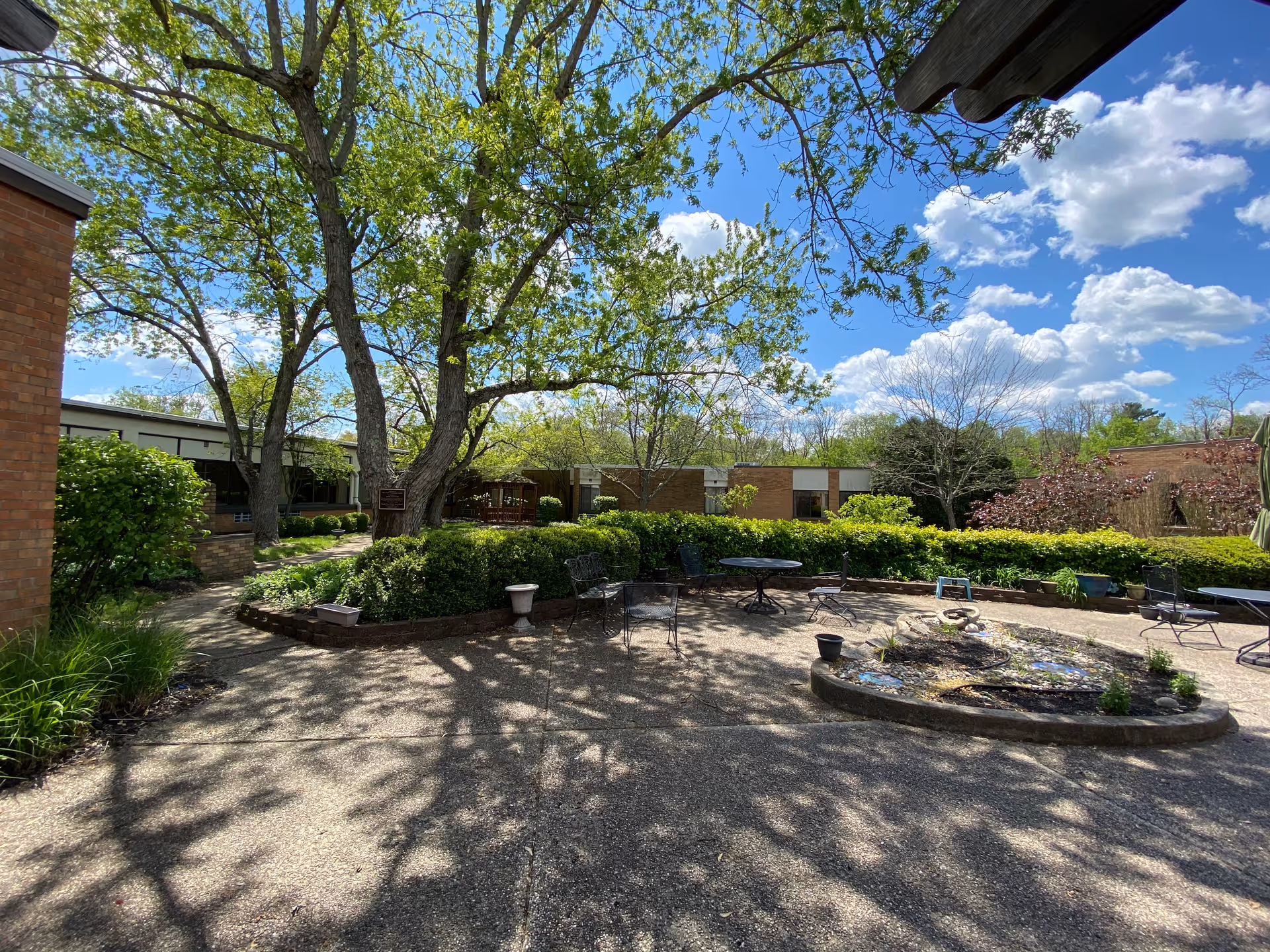 Outdoor courtyard area at Alois Alzheimer Center with trees, shrubs, and patio furniture including tables and chairs. The sky is blue with scattered clouds, and the area is surrounded by single-story brick buildings.