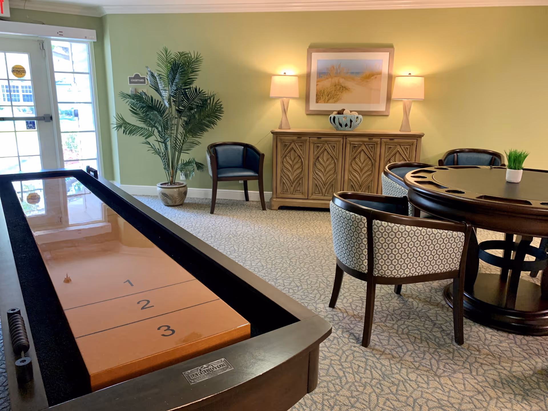 A cozy indoor recreational room with a shuffleboard table in the foreground, a round table with four patterned chairs, a wooden cabinet with two lamps and a framed beach-themed picture above it, a potted plant near a glass door leading outside, and carpeted flooring.