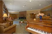A spacious and well-lit living room area in a senior living facility featuring a grand piano with a vase of flowers on top, comfortable armchairs, a coffee table, a fireplace, and large windows with curtains allowing natural light to fill the room.