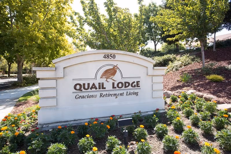 Stone entrance sign reading "Quail Lodge Gracious Retirement Living" with the address 4850, set among flowers and trees.