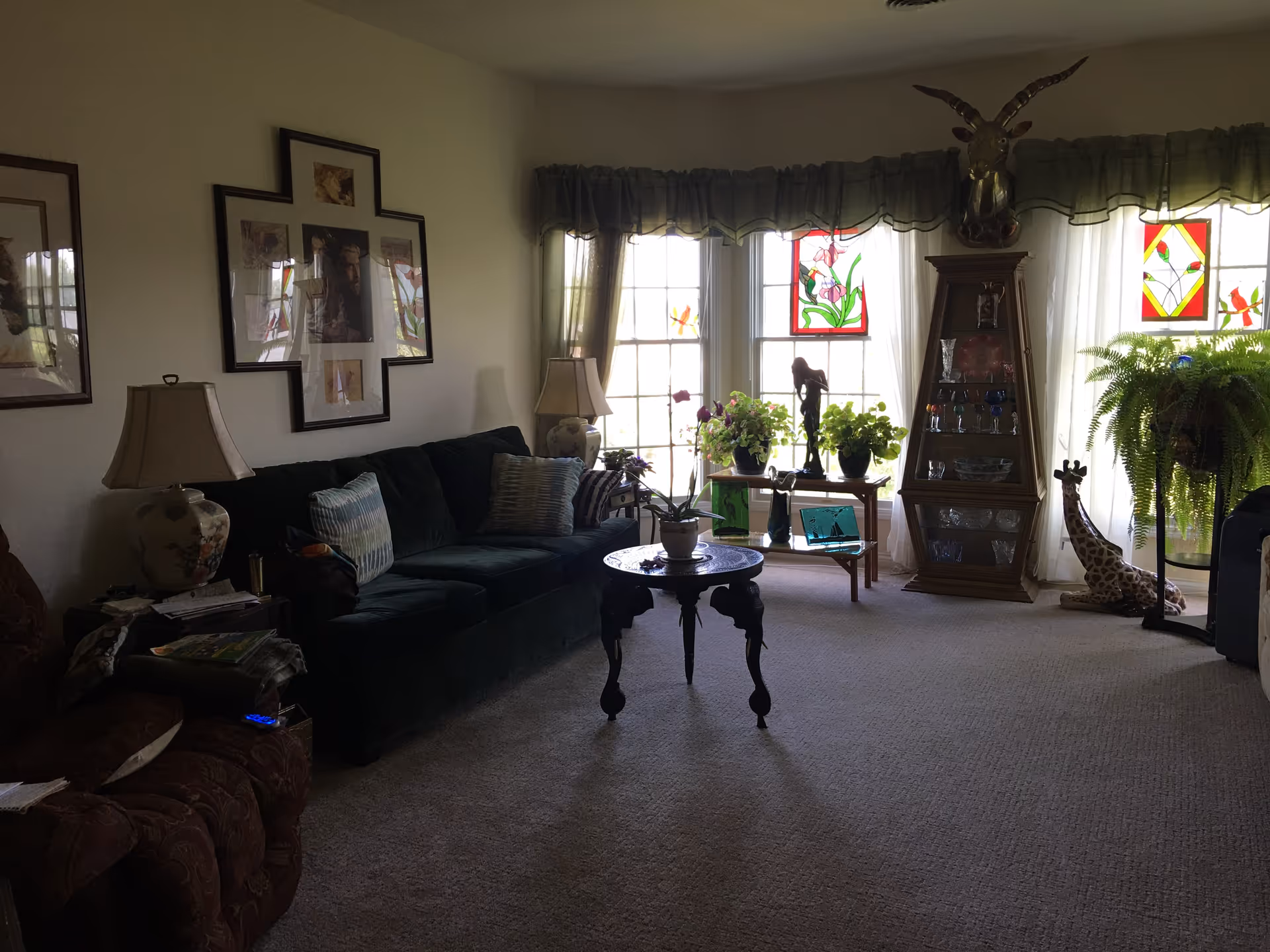 A living room with a dark green couch and a brown armchair. There are two table lamps on either side of the couch. The room has a carpeted floor and large windows with sheer curtains and stained glass panels. Several potted plants are placed on a small table in front of the windows. A wooden display cabinet with glassware and a decorative animal head mounted above it is visible. A giraffe figurine and a large fern plant are also present near the windows.