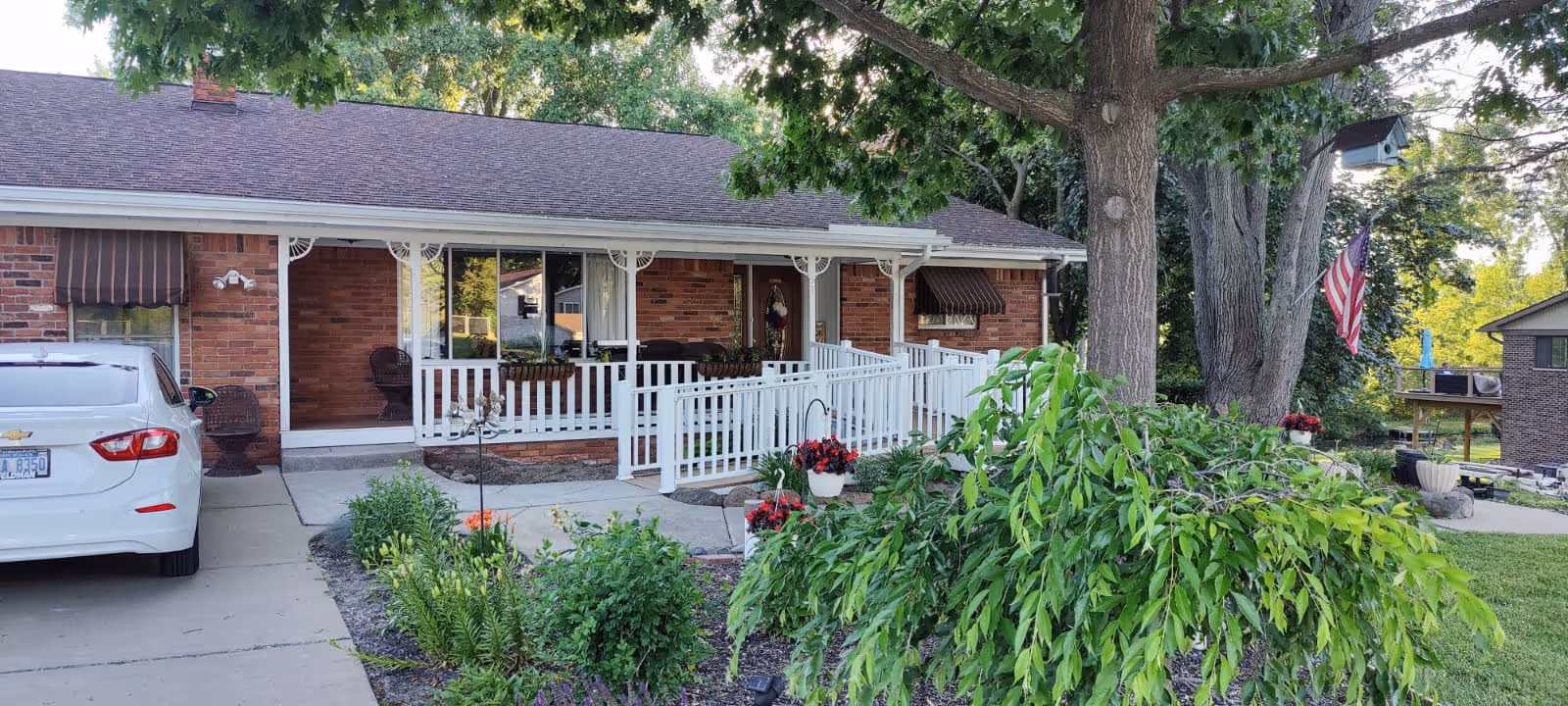 Front of a single-story brick home with a covered porch, white accessibility ramp and railings, landscaped yard, parked car and an American flag.