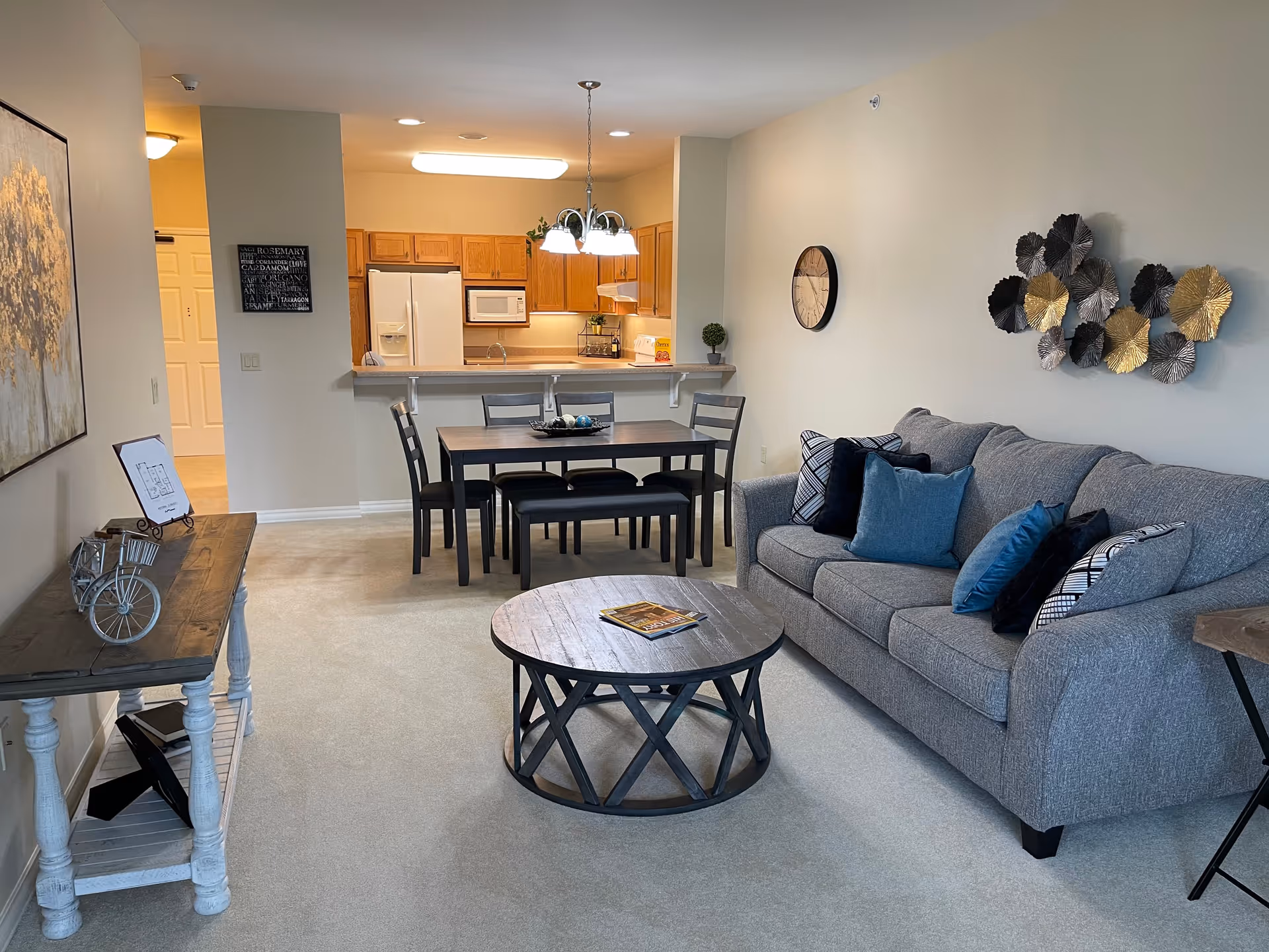 A cozy living and dining area in a senior living facility apartment. The room features a gray sofa with multiple decorative pillows, a round wooden coffee table with a magazine on top, and a dining table with four chairs and a bench. The kitchen is visible in the background with wooden cabinets, a white refrigerator, microwave, and stove. The walls are decorated with a clock and metal wall art, and the floor is carpeted.