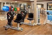 Interior view of a fitness room in a senior living facility featuring exercise equipment including a seated leg press machine, colorful exercise balls on a rack, a rack of dumbbells, and a television mounted on the wall. The room has wooden flooring and large mirrors on the walls.