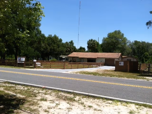 View of a single-story building with a reddish-brown roof behind a wooden fence, surrounded by trees and greenery, with a paved road in the foreground under a clear blue sky.