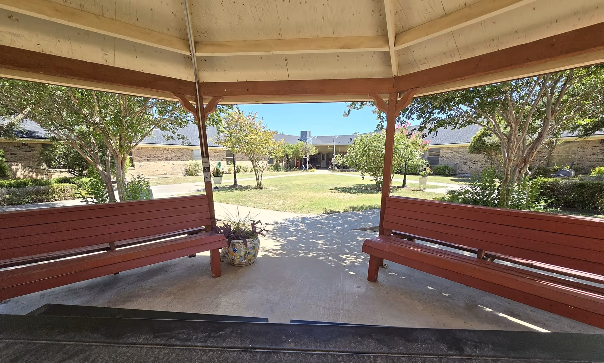 View from inside a wooden gazebo with red benches on both sides, overlooking a courtyard with green grass, trees, and a building in the background under a clear blue sky.