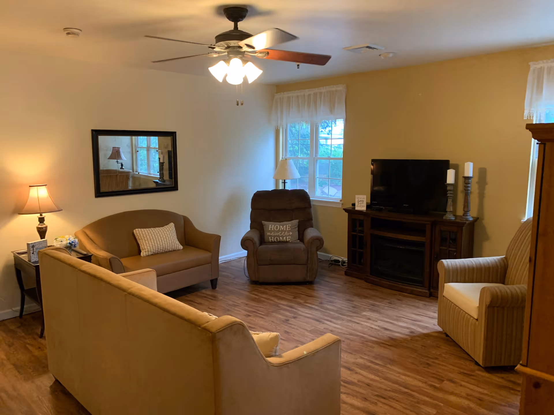 A cozy living room with wooden flooring, featuring a beige sofa, a brown loveseat with a white pillow, a brown recliner with a pillow that says 'Home Sweet Home', and a beige striped armchair. There is a wooden TV stand with a flat-screen TV and two candle holders. A ceiling fan with lights is on the ceiling, and there are two windows with white curtains letting in natural light. A table lamp is on a side table next to the loveseat, and a rectangular mirror hangs on the wall above it.