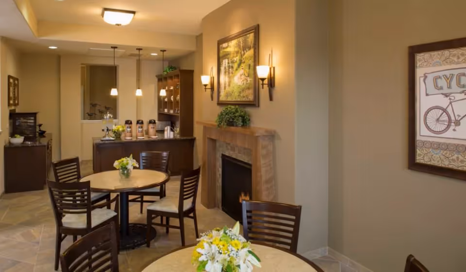 A cozy dining area with round tables and wooden chairs, each table decorated with a vase of yellow and white flowers. The room features a fireplace with a framed nature painting above it, warm wall sconces, and a counter in the background with coffee dispensers and hanging pendant lights.