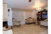 Carpeted living room with traditional wooden-framed sofas and armchairs, a TV in a cabinet, chandelier, and large curtained window.