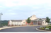 Exterior view of a large, beige-colored nursing center building with a covered entrance, surrounded by a paved driveway and landscaped greenery under a cloudy sky.