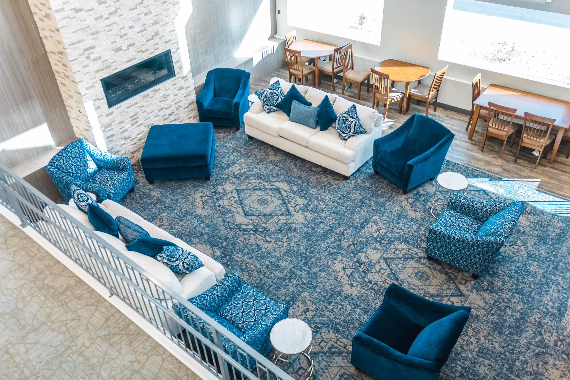 A bright and spacious living room area in Downtown Assisted Living featuring two white sofas with blue patterned and solid blue pillows, several blue armchairs, a large blue patterned rug, a modern fireplace with a stone surround, and wooden tables with chairs near large windows letting in natural light.