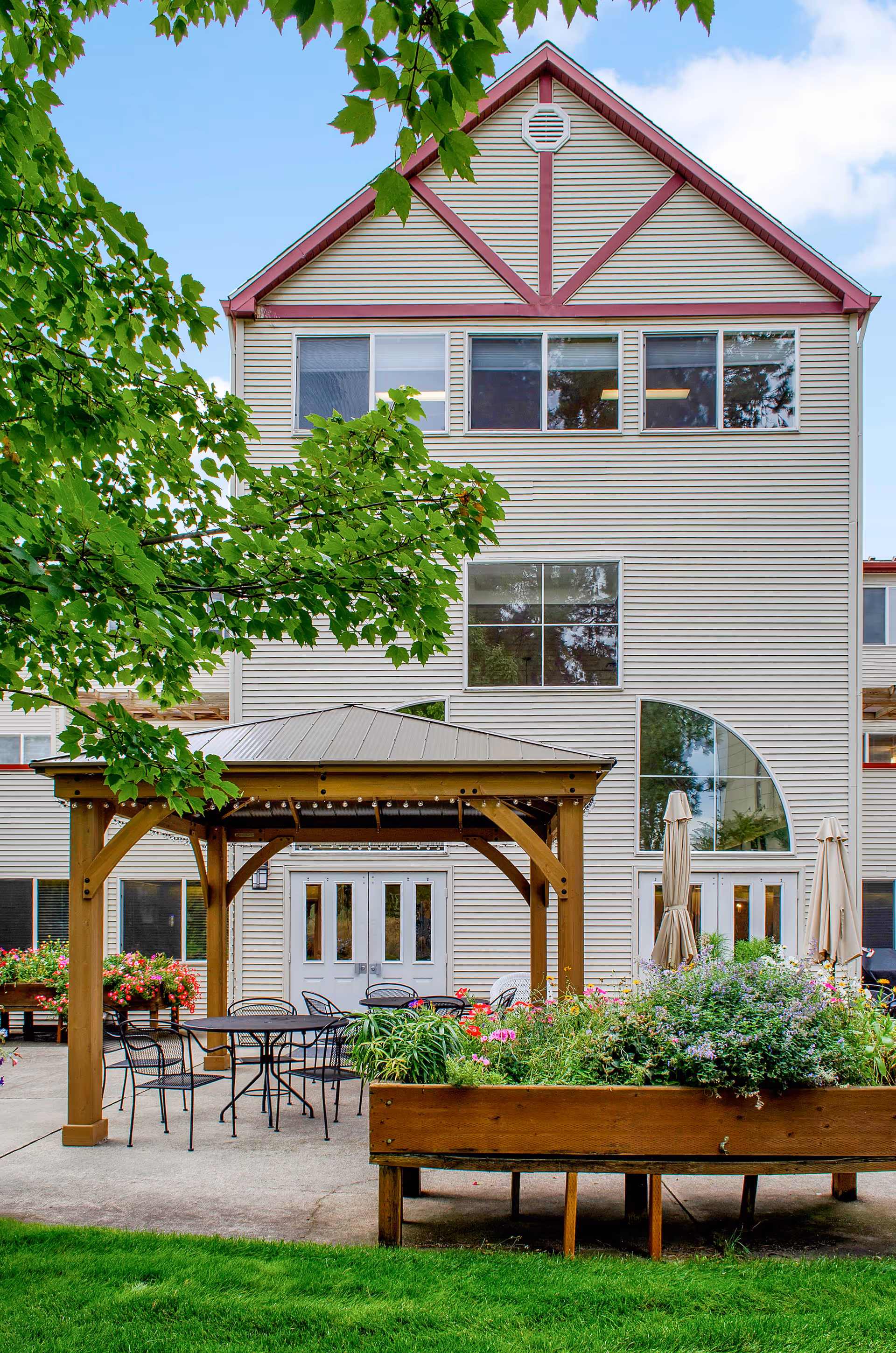 Outdoor patio area at North Star Assisted & Senior Living featuring a wooden gazebo with metal roof, several black metal tables and chairs, surrounded by flower beds and greenery, with the multi-story building facade in the background.