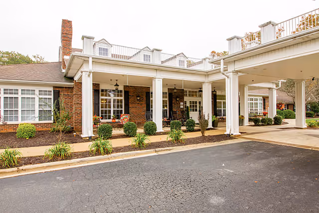 Exterior view of Brookdale Lawndale Park showing a brick building with white columns supporting a covered entrance. There are windows with white frames, a chimney, and landscaping with bushes and plants along the walkway.