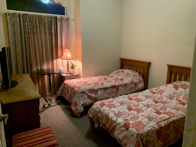 Small bedroom with two twin beds covered in red floral bedspreads, a bedside table with a lamp, a TV on a dresser, and curtains over the window.
