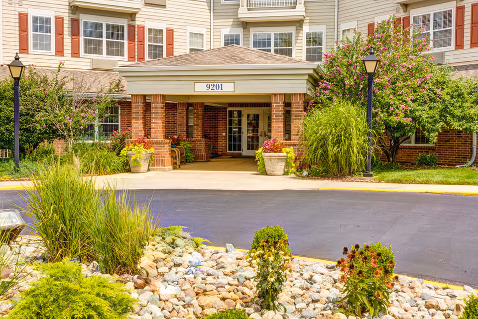 Covered entrance and driveway of a senior living building with '9201' sign, brick columns, and landscaped plants.