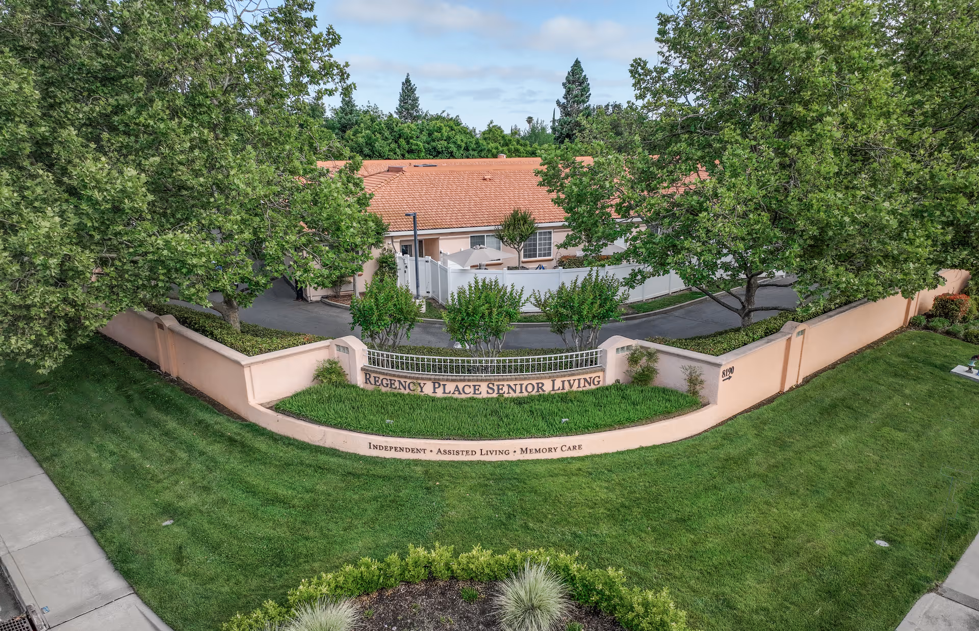 Exterior view of Regency Place Senior Living facility showing a peach-colored wall with the facility name and services offered, surrounded by green grass, trees, and a sidewalk.